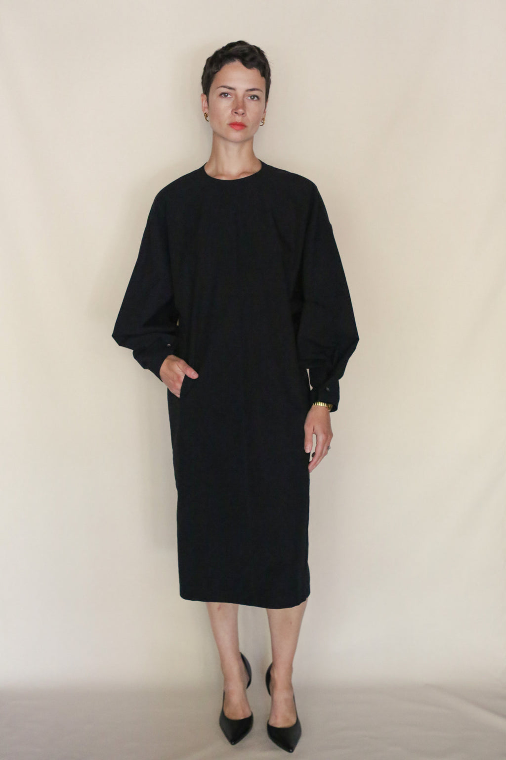A person with short dark hair wears the Cocoon Dress | Black with black pointed-toe heels, standing against a plain beige background with one hand in their pocket. Handmade San Francisco craftsmanship is evident.