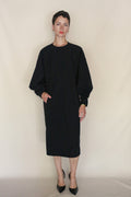 A person with short dark hair wears the Cocoon Dress | Black with black pointed-toe heels, standing against a plain beige background with one hand in their pocket. Handmade San Francisco craftsmanship is evident.