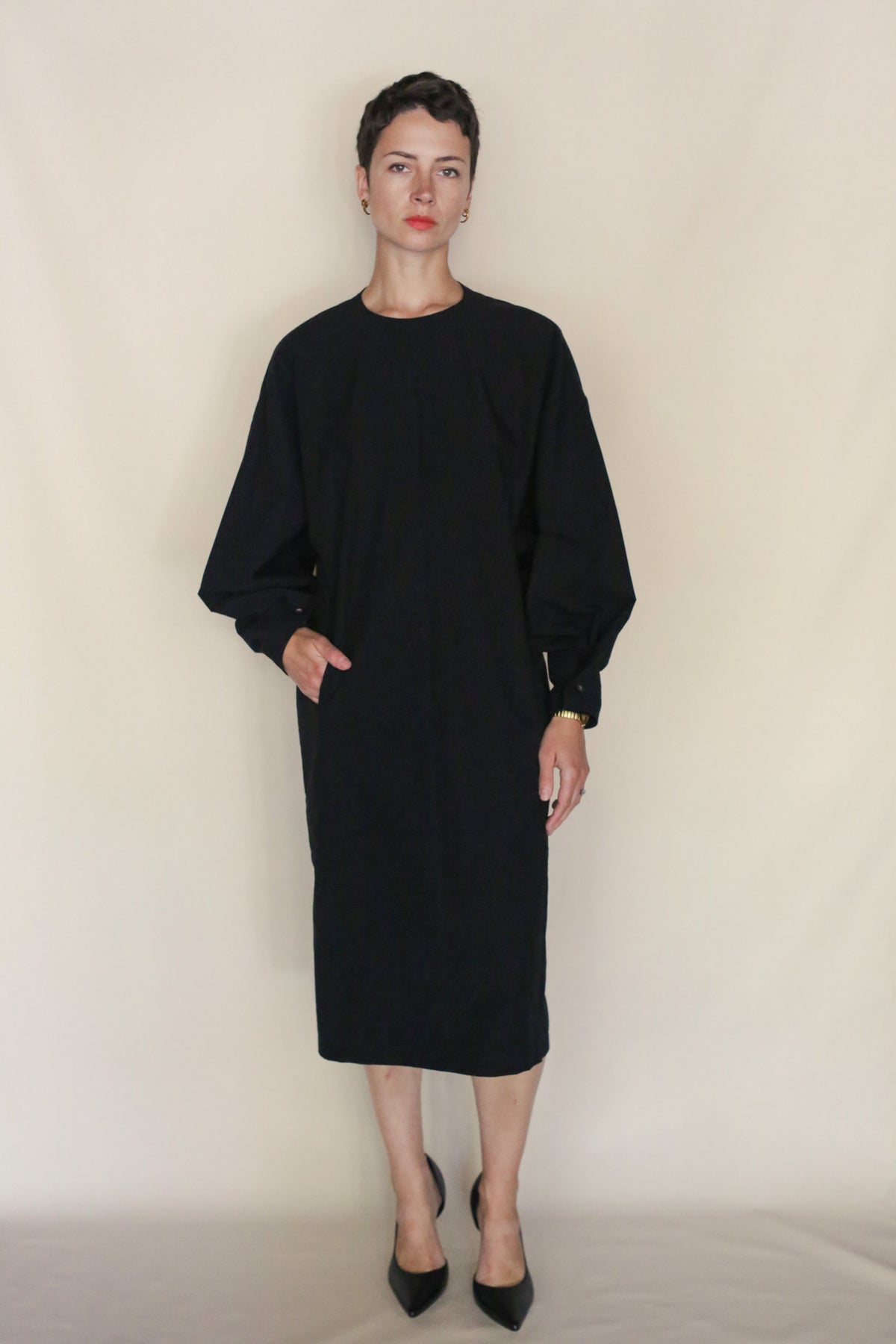 A person with short dark hair wears the Cocoon Dress | Black with black pointed-toe heels, standing against a plain beige background with one hand in their pocket. Handmade San Francisco craftsmanship is evident.
