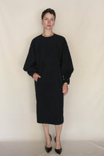 Load image into Gallery viewer, A person with short dark hair wears the Cocoon Dress | Organic Typewriter Cotton Black, handmade in San Francisco, paired with black heels, standing against a plain beige background with one hand in their pocket.