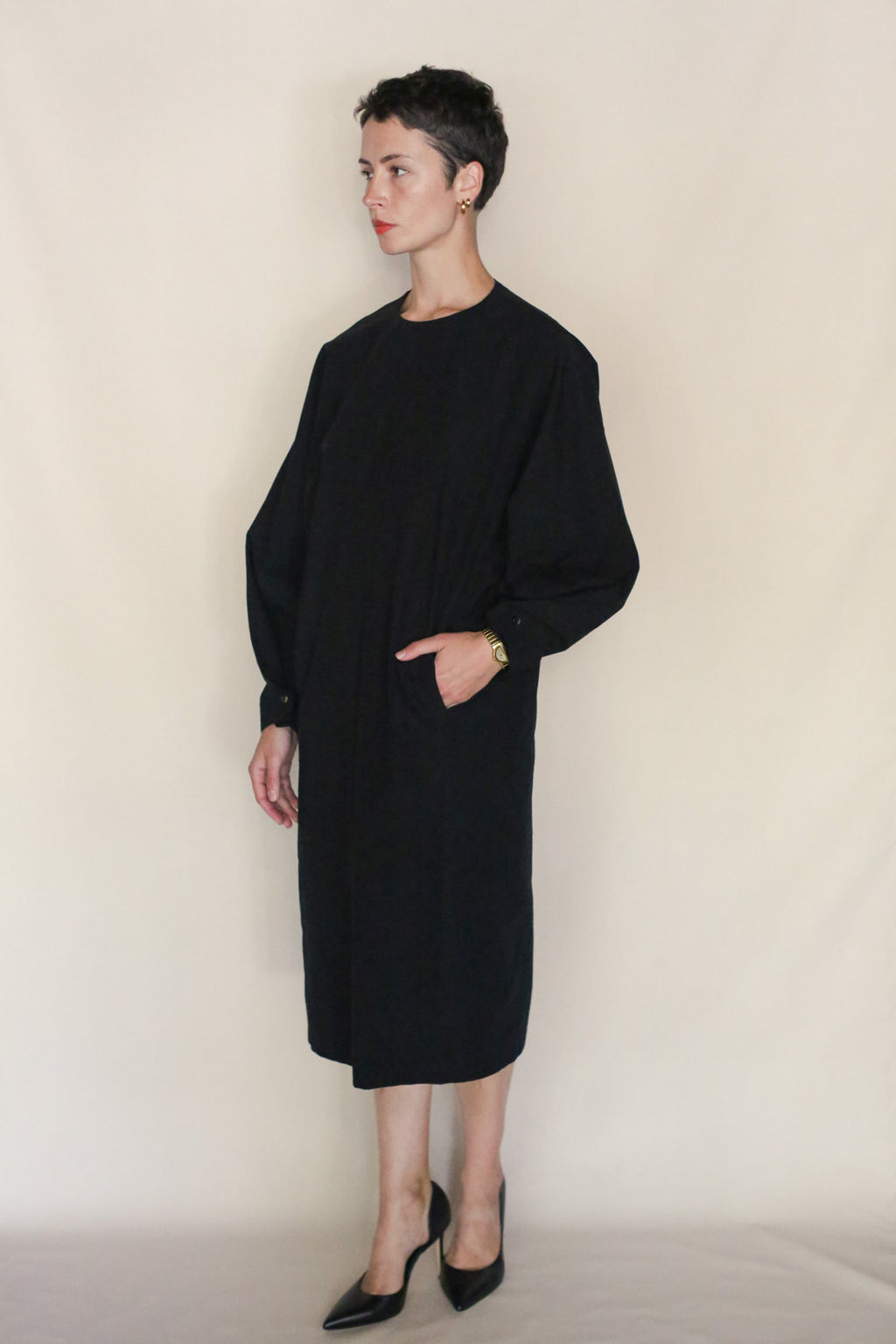 A person with short hair wears the Cocoon Dress | Black—loose-fitting, long-sleeved, and made from Organic Typewriter Cotton—with black heels and a hand in their pocket, standing against a plain beige background. This look reflects Handmade San Francisco style.