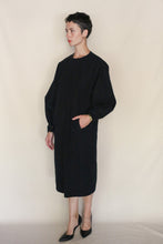 Load image into Gallery viewer, A person with short hair wears the Cocoon Dress | Organic Typewriter Cotton Black, a loose-fitting, long-sleeved piece, standing against a beige background with one hand in a pocket, showcasing Handmade San Francisco style.