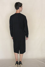 Load image into Gallery viewer, A person with short hair, wearing the Cocoon Dress | Organic Typewriter Cotton Black and black heels, stands facing away from the camera against a plain light background. Handmade in San Francisco.