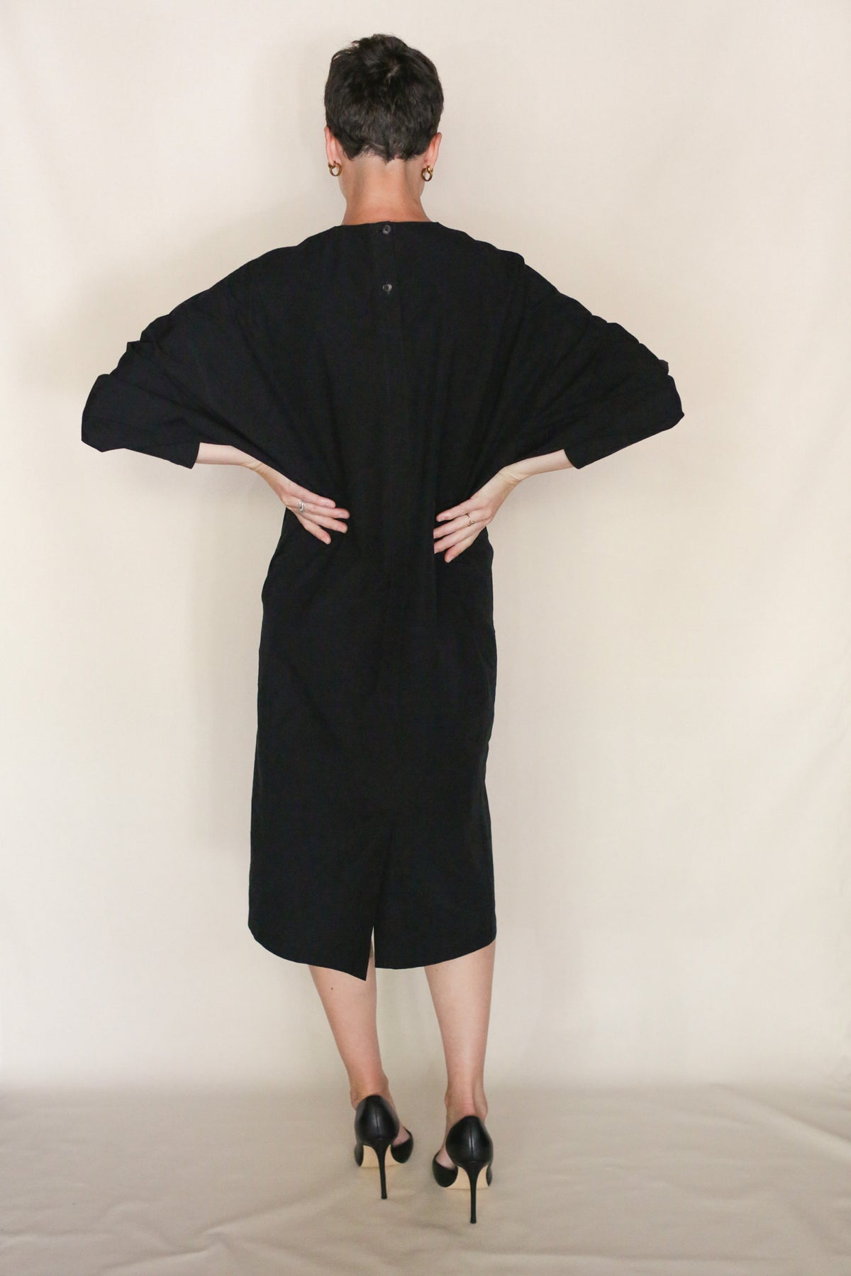 A person with short hair stands facing away, wearing the loose-fitting Cocoon Dress | Black with long sleeves and black heels, hands on hips against a plain light background. Handmade San Francisco craftsmanship is evident.