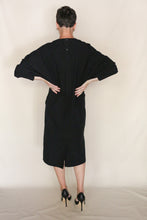 Load image into Gallery viewer, A short-haired person, wearing the Cocoon Dress | Organic Typewriter Cotton Black and black high heels, stands with their back to the camera and hands on hips against a plain light-colored background.