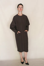 Load image into Gallery viewer, A person with short dark hair wears the Cocoon Dress | Organic Typewriter Cotton Earth—handmade in San Francisco with pockets—paired with black pointed-toe heels, standing against a plain light background.