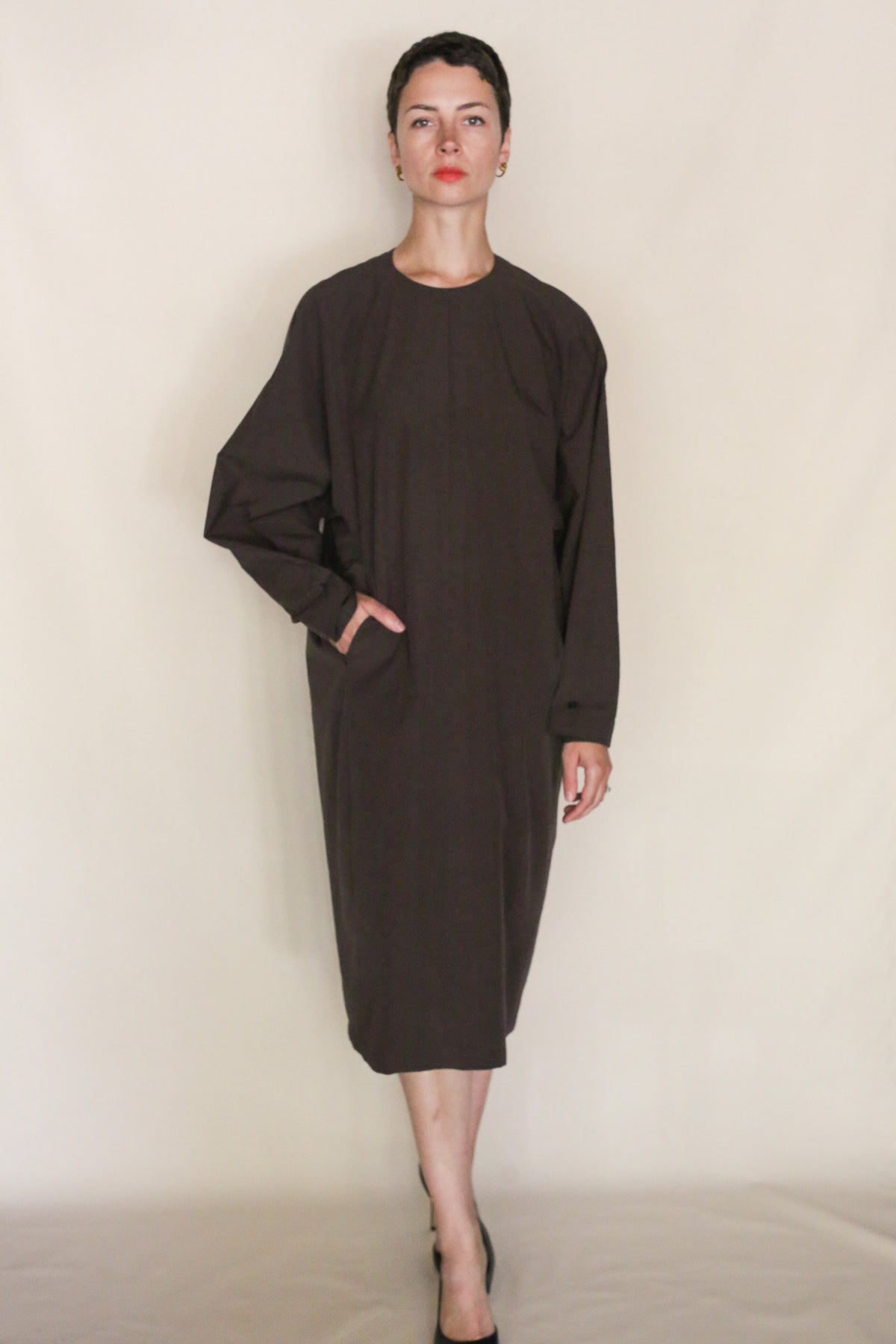 A person with short dark hair and black shoes stands against a plain beige background, wearing the loose-fitting, long-sleeved Cocoon Dress | Earth made from Organic Typewriter Cotton, with one hand in a pocket and a neutral expression.