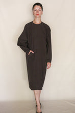 Load image into Gallery viewer, A person with short dark hair wears the Cocoon Dress | Organic Typewriter Cotton Earth—a loose, long-sleeved dress—with black shoes, standing against a plain beige background and one hand in a pocket.