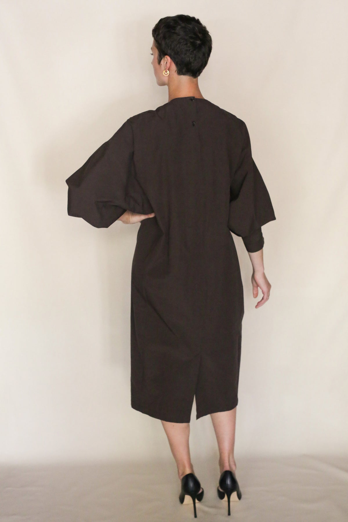 A person with short dark hair, seen from behind, wears the loose-fitting handmade Cocoon Dress | Earth crafted from organic typewriter cotton in dark brown, paired with black heels against a plain neutral background.