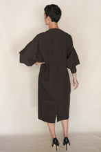 Load image into Gallery viewer, A person with short dark hair stands with their back to the camera, wearing the Cocoon Dress | Organic Typewriter Cotton Earth and black high heels against a plain, light background.