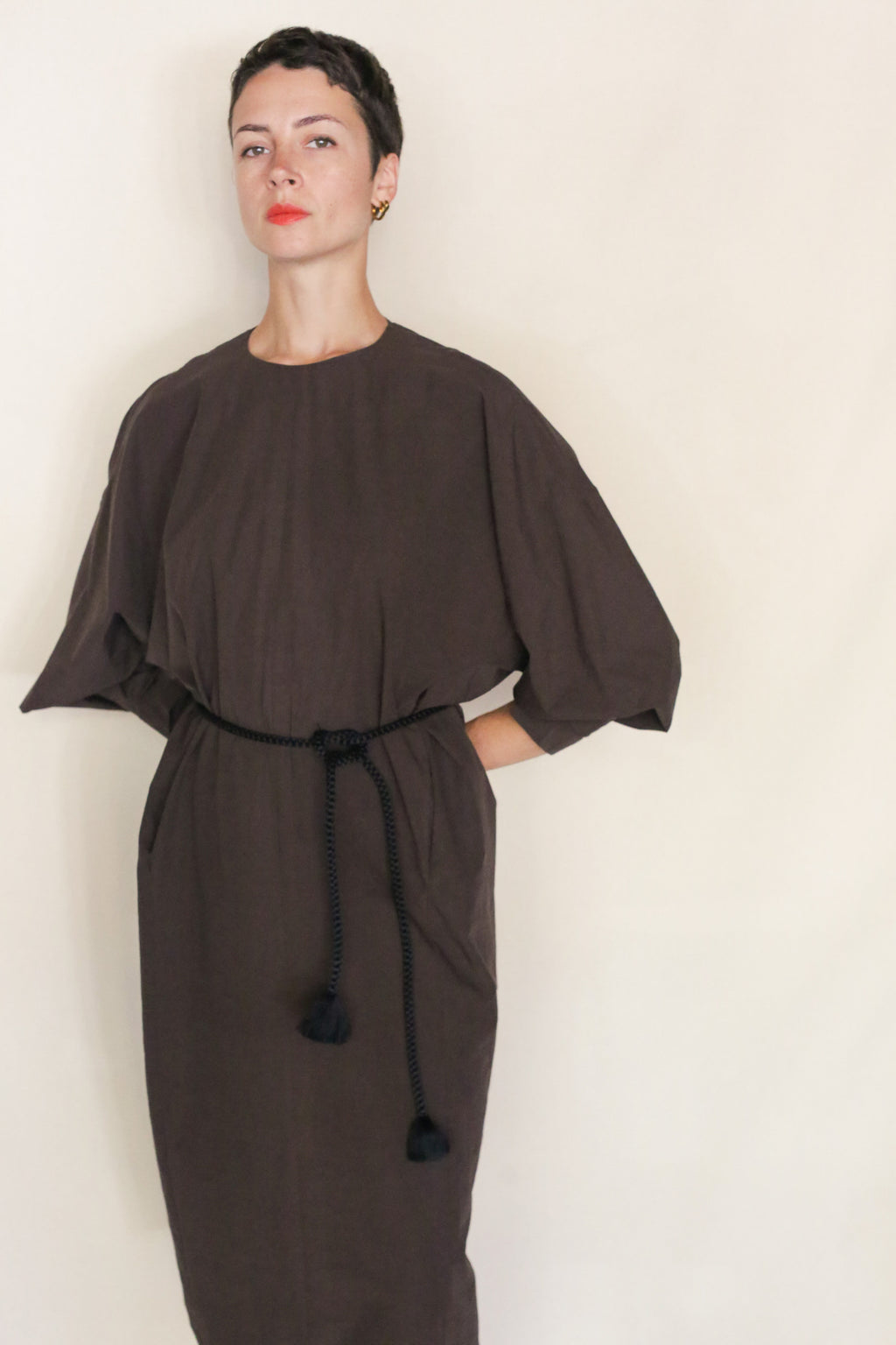 A person with short dark hair wears the Cocoon Dress | Earth, a loose-fitting, dark brown dress of organic typewriter cotton with a black rope belt and wide sleeves, handmade in San Francisco, against a plain light background.