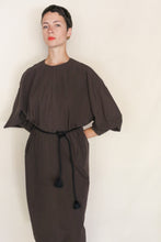 Load image into Gallery viewer, A person with short dark hair wears the Cocoon Dress | Organic Typewriter Cotton Earth—loose-fitting, dark brown with wide sleeves and a black rope belt, handmade in San Francisco—standing against a plain light background.