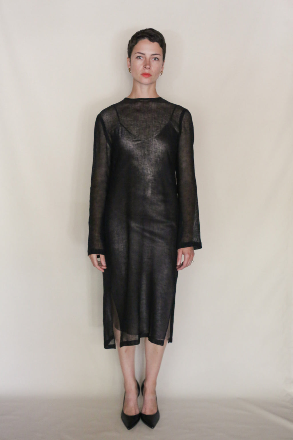 A person with short hair faces forward against a plain, light background. They wear the Leno Crewneck Dress in sheer black linen and black heels, maintaining a neutral facial expression.
