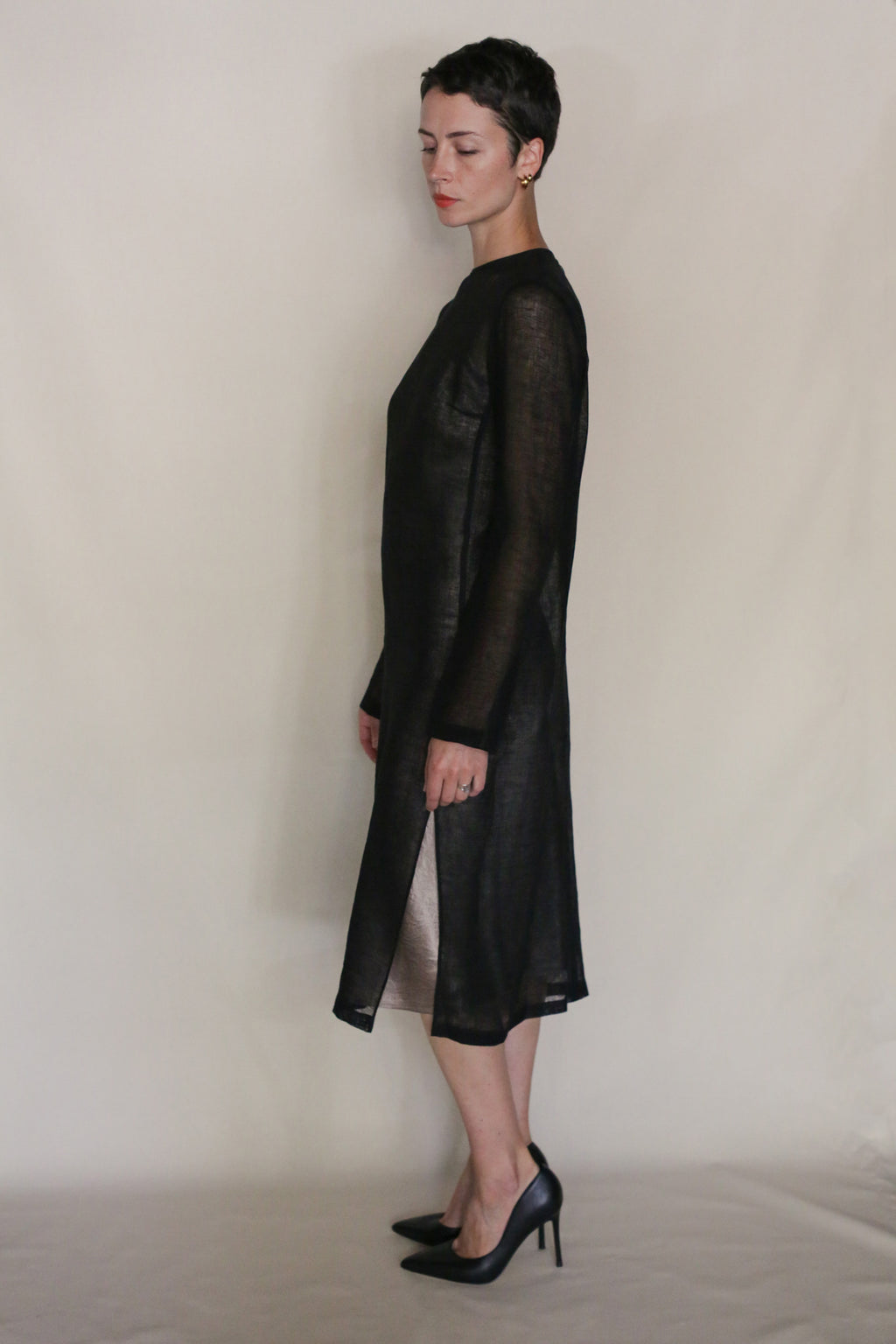 A person with short dark hair stands in profile, wearing a sheer black Leno Crewneck Dress and black high heels against a plain light background—a chic, handmade San Francisco look.