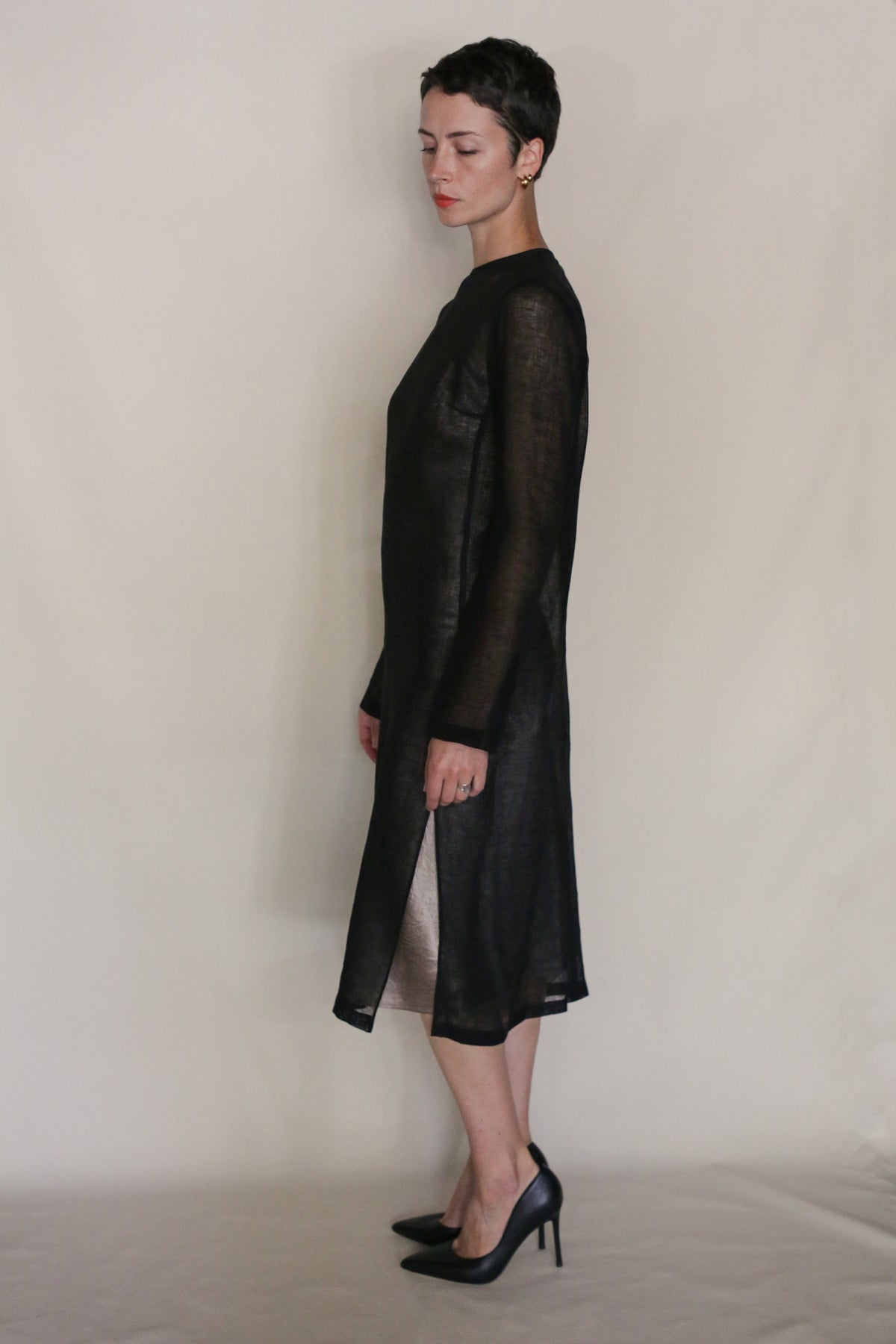 A person with short dark hair stands in profile, wearing a sheer black Leno Crewneck Dress and black high heels against a plain light background—a chic, handmade San Francisco look.