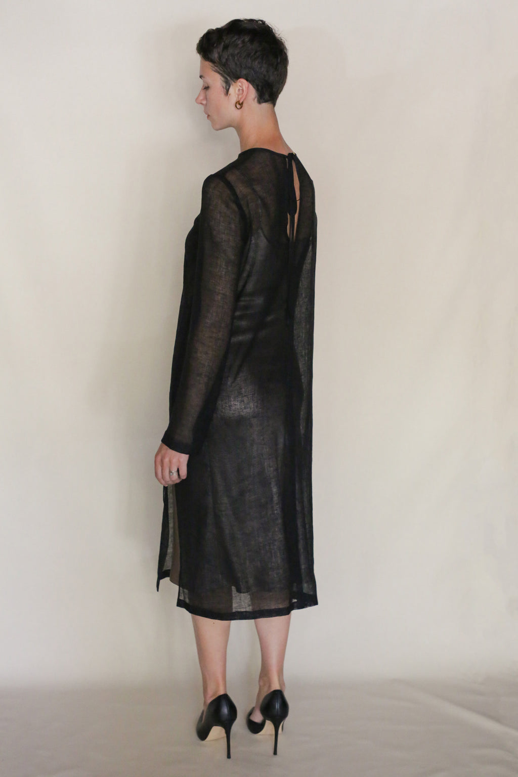 A person with short dark hair, wearing the Leno Crewneck Dress in sheer black with long sleeves and black high heels, stands facing away against a plain, light background.