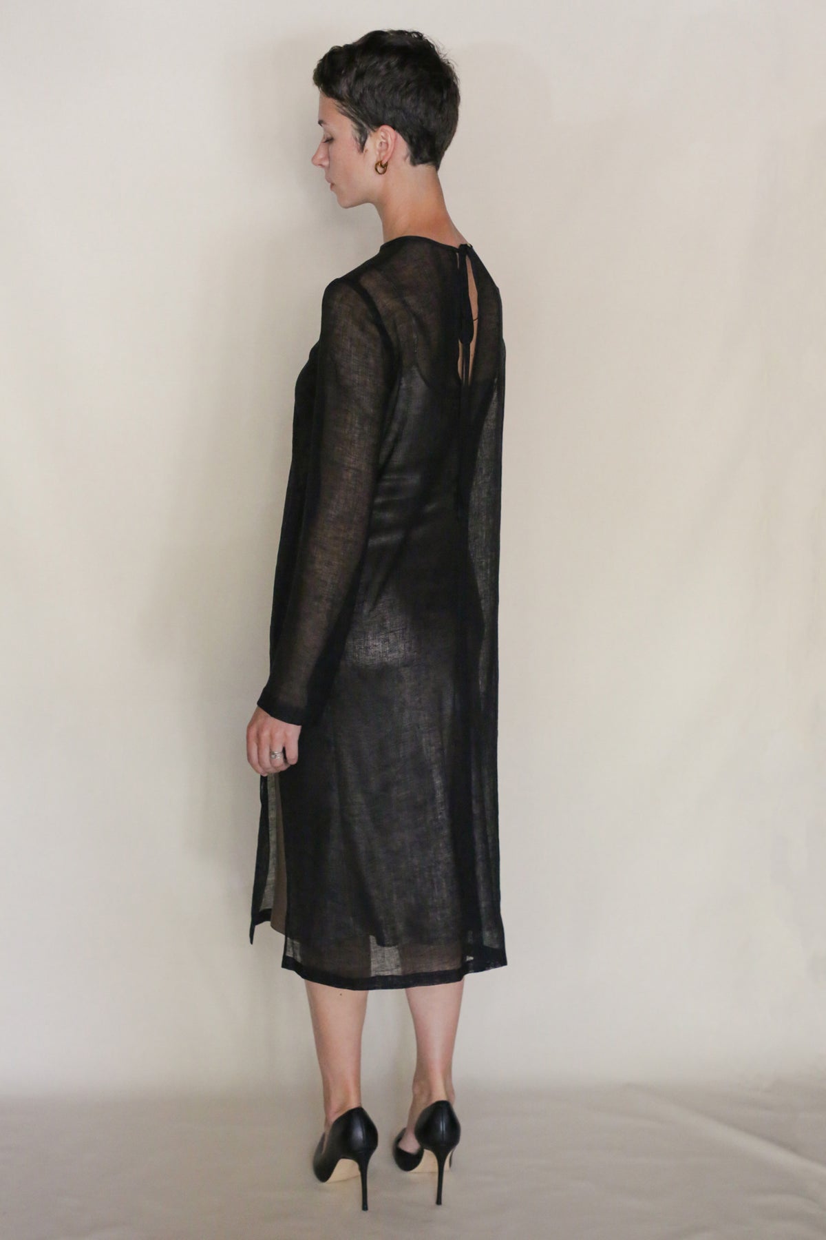A person with short dark hair, wearing the Leno Crewneck Dress in sheer black with long sleeves and black high heels, stands facing away against a plain, light background.