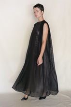 Load image into Gallery viewer, A person with short dark hair models the Shoulder Pleat Dress | Linen Leno Black—a sleeveless, floor-length, sheer black dress—paired with black heels against a light background, embodying modern elegance inspired by handmade San Francisco design.