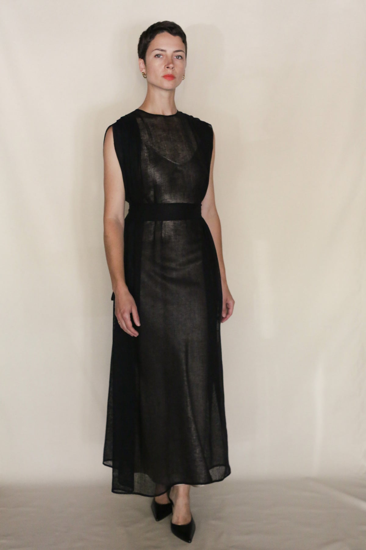 A woman with short dark hair wears the Leno Pleat Dress—a sleeveless, sheer black ankle-length dress with a subtle shoulder pleat and sash belt—paired with black heels, standing against a plain light background channeling handmade San Francisco style.