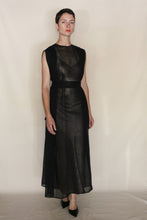 Load image into Gallery viewer, A person with short dark hair wears the Shoulder Pleat Dress | Linen Leno Black, a sleeveless, sheer black dress with pleated shoulders and a waist sash, paired with black heels against a plain light background.
