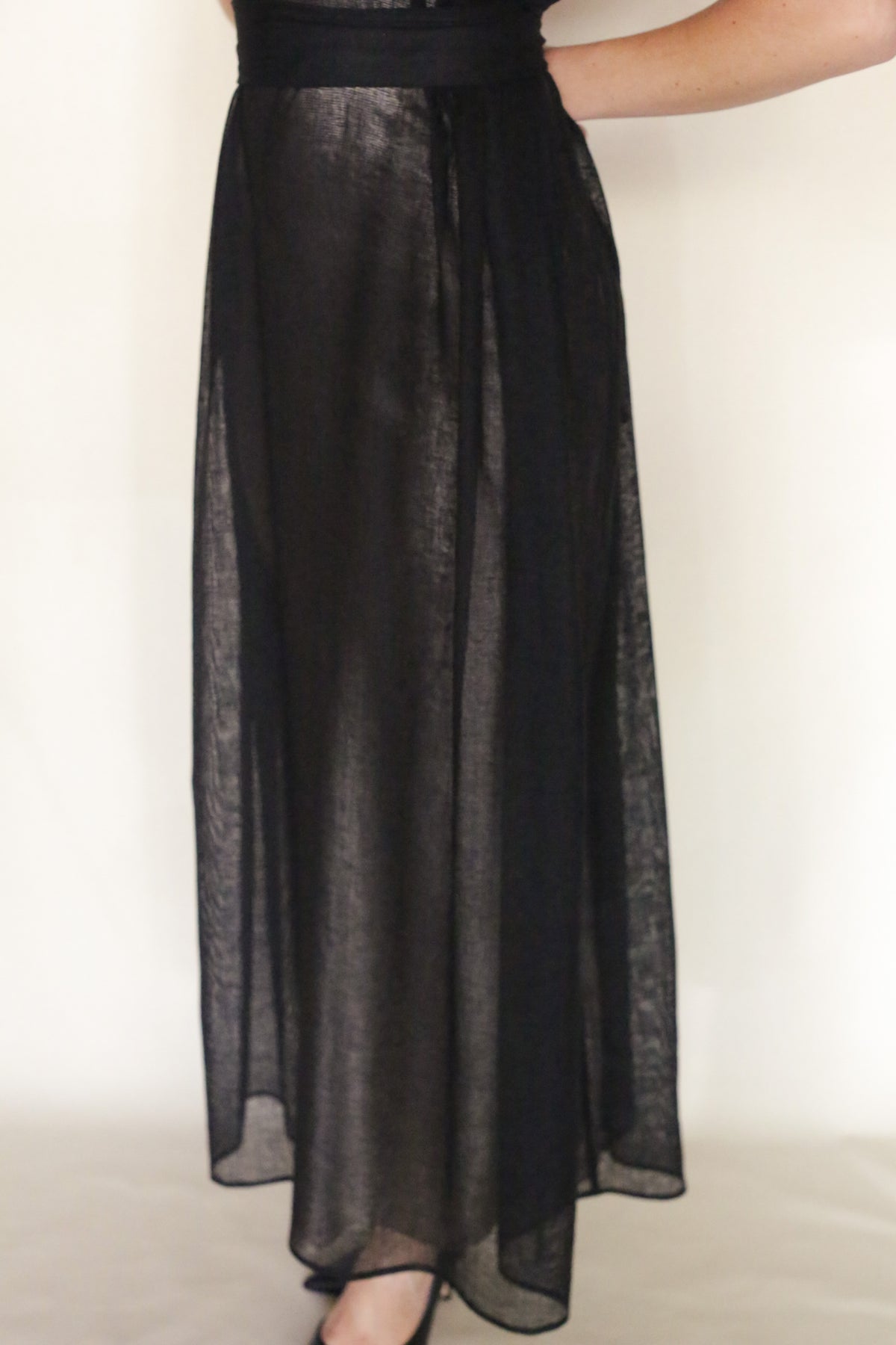 A person models the Leno Pleat Dress—a sheer black maxi skirt with a black underskirt and subtle shoulder pleat—against a plain light background, showing only the lower body and part of an arm.