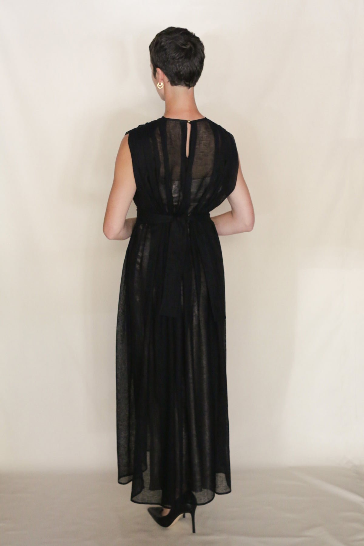 A person with short dark hair, wearing the Leno Pleat Dress—a sleeveless, sheer black linen dress with subtle shoulder pleats—and black high heels stands facing away from the camera against a plain light background.