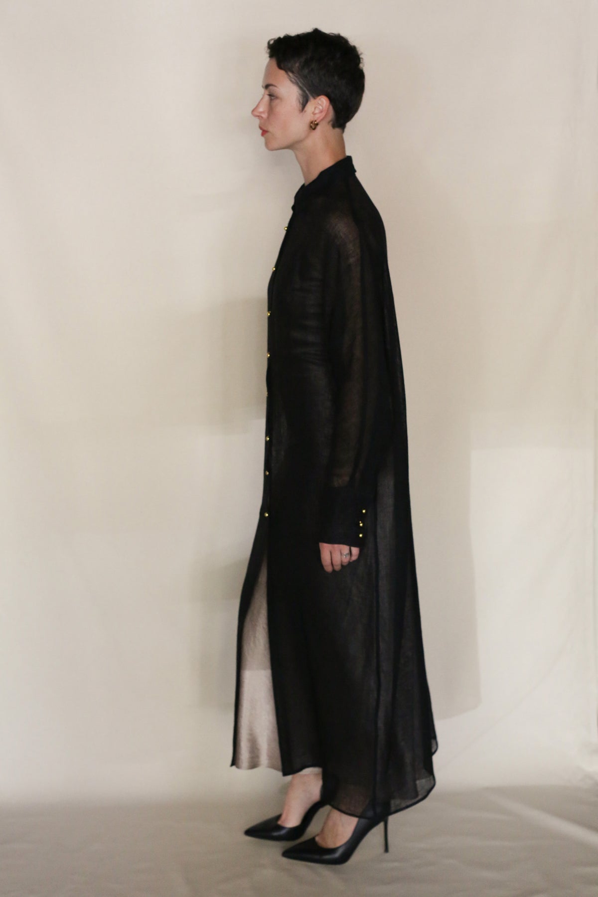 A person with short dark hair stands in profile against a plain background, wearing the Leno Dresscoat by Atelier Lan Jaenicke—a long, sheer black coat handcrafted in San Francisco—paired with black high-heeled shoes.