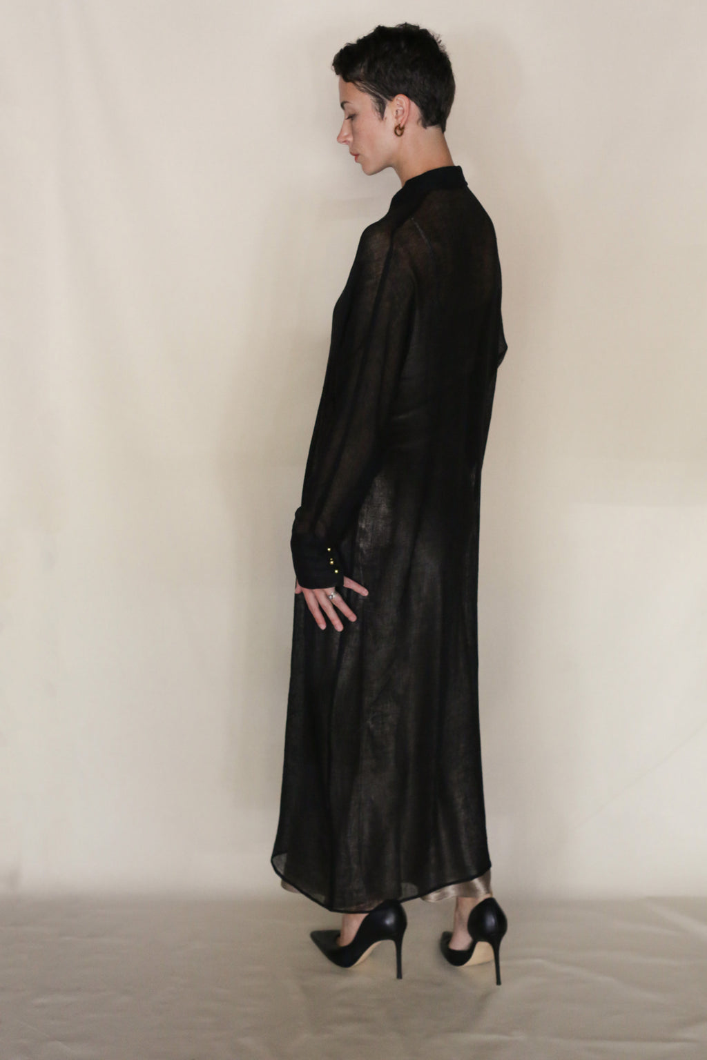 A person with short dark hair stands sideways in a sheer black long-sleeve dress, black heels, and the Leno Dresscoat by Atelier Lan Jaenicke, set against a plain light background.