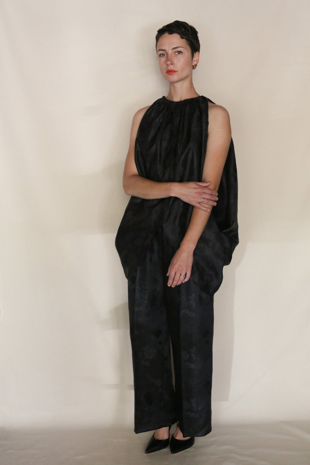 Wearing the Lingering Fragrance Butterfly, a person with short dark hair poses against a light background in a loose, sleeveless black silk jacquard outfit with wide legs and gathered neckline, paired with black pointed shoes and arms softly crossed.