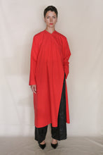 Load image into Gallery viewer, A person with short dark hair and a neutral expression wears the Qipao Dresscoat | Organic Typewriter Vermilion over black wide-leg pants and pointed shoes, standing against a plain backdrop.