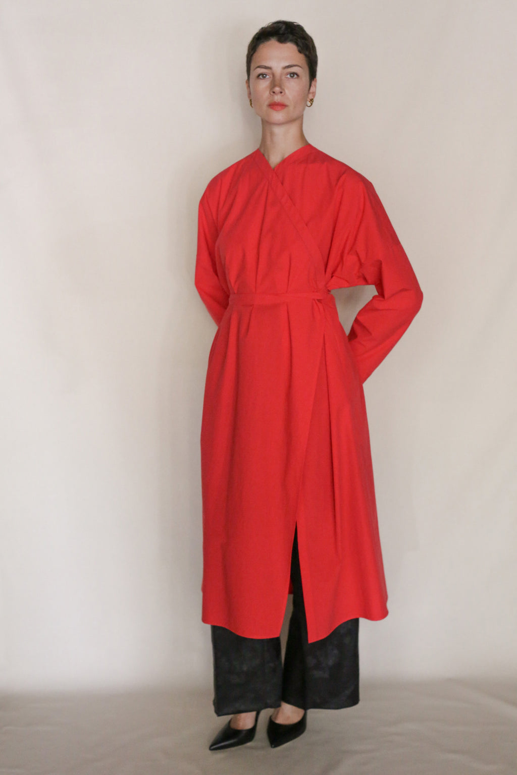 A person stands against a plain background wearing the handmade Qipao Dresscoat | Vermilion with long sleeves and a side slit, layered over wide-legged black pants and black pointed-toe shoes.