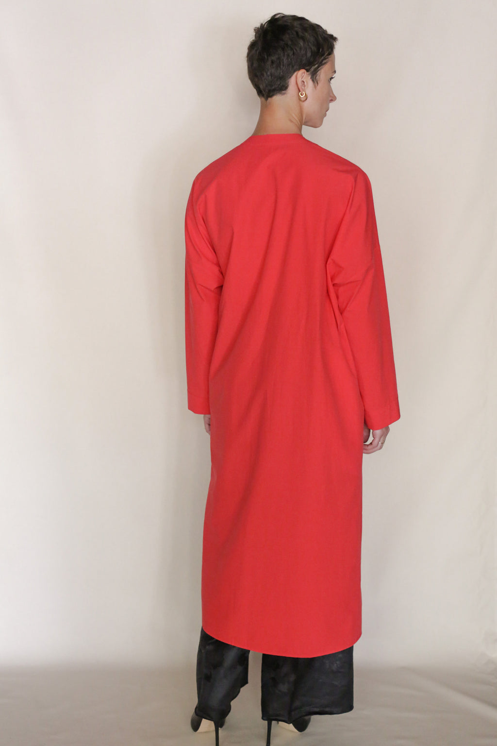 A person stands facing away, wearing the Qipao Dresscoat | Vermilion— a long, loose-fitting red coat made from organic typewriter cotton—over black pants and shoes against a plain light background. Handmade in San Francisco.