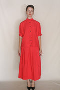 A person stands against a plain backdrop wearing a bright red Organic Typewriter Cotton button-up shirt with rolled sleeves and the Column Wrap Skirt | Vermillion, paired with black heels for a chic, zero-waste design look.