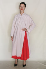 Load image into Gallery viewer, A person with short dark hair and a neutral expression wears a loose, light pink long-sleeved dress over the Column Wrap Skirt | Organic Typewriter Cotton Vermillion, paired with black pointed heels against a light background.