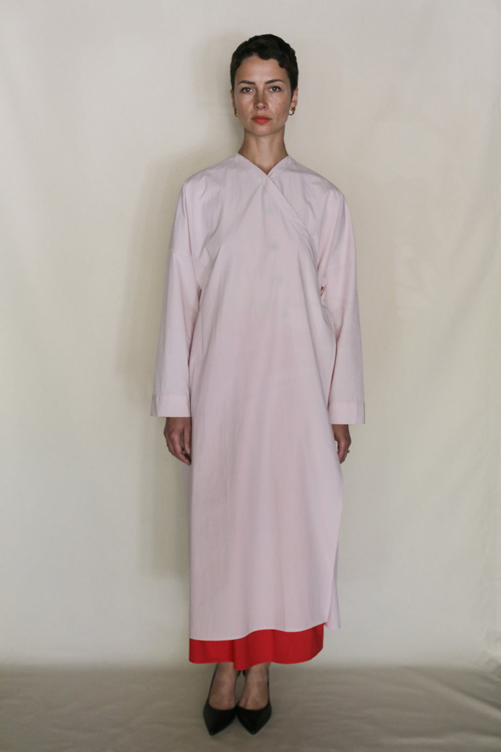 A person stands against a plain light background, wearing the Qipao Dresscoat | Blush— a long, loose pale pink coat made from organic typewriter cotton with a red hem showing. They have short dark hair and wear black pointed shoes, exuding handmade San Francisco style.