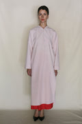 A person stands against a plain light background, wearing the Qipao Dresscoat | Blush— a long, loose pale pink coat made from organic typewriter cotton with a red hem showing. They have short dark hair and wear black pointed shoes, exuding handmade San Francisco style.