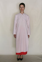 Load image into Gallery viewer, A person stands against a plain background wearing the Qipao Dresscoat | Organic Typewriter Cotton Blush, a long, loose-fitting light pink dress coat with long sleeves and a red hem, paired with black heels. Handmade in San Francisco.