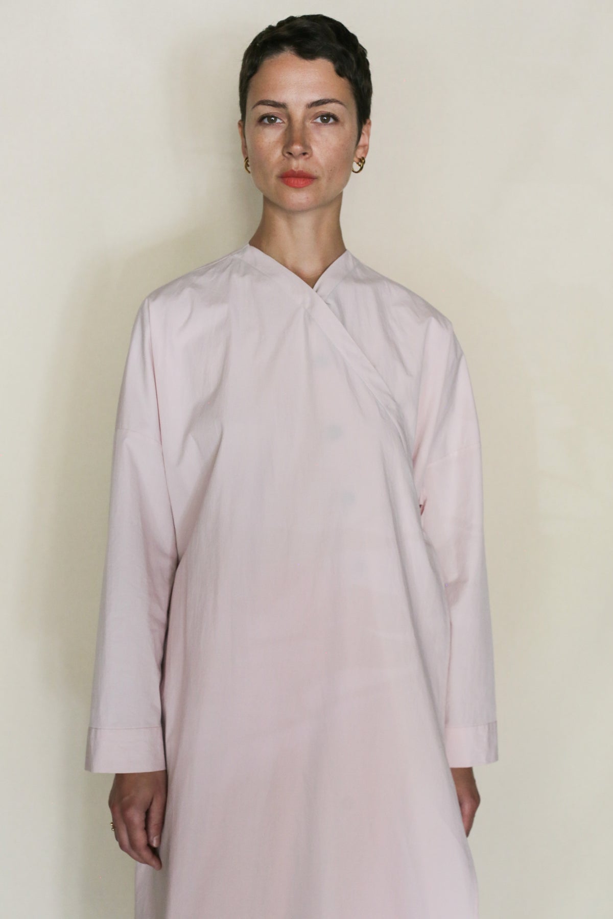 A person with short dark hair, wearing the Qipao Dresscoat | Blush, stands against a plain, light background and looks directly at the camera with a neutral expression.