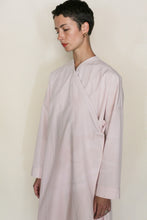Load image into Gallery viewer, A person with short dark hair wears the Qipao Dresscoat in Organic Typewriter Cotton Blush, featuring a loose, long-sleeved wrap design, standing against a plain beige background and looking down with a calm expression.