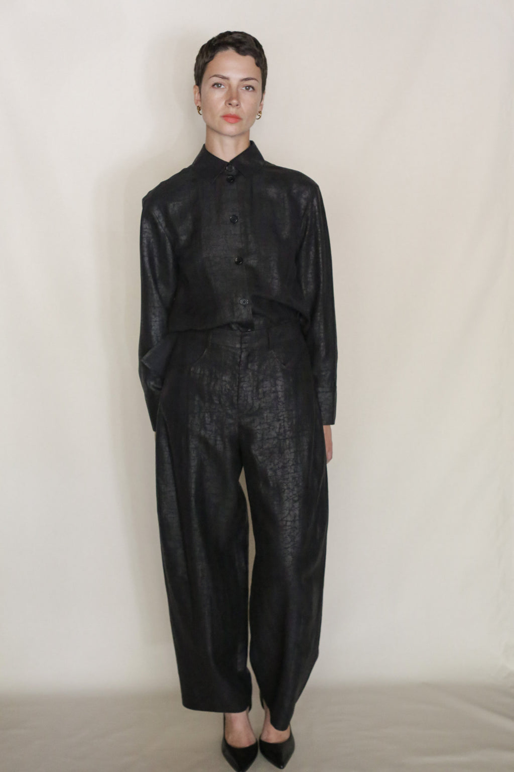 A person with short hair stands against a plain light background, wearing a loose-fitting, shiny black Earth Mark Studio Shirt tucked into matching high-waisted, wide-leg pants and black pointed shoes.