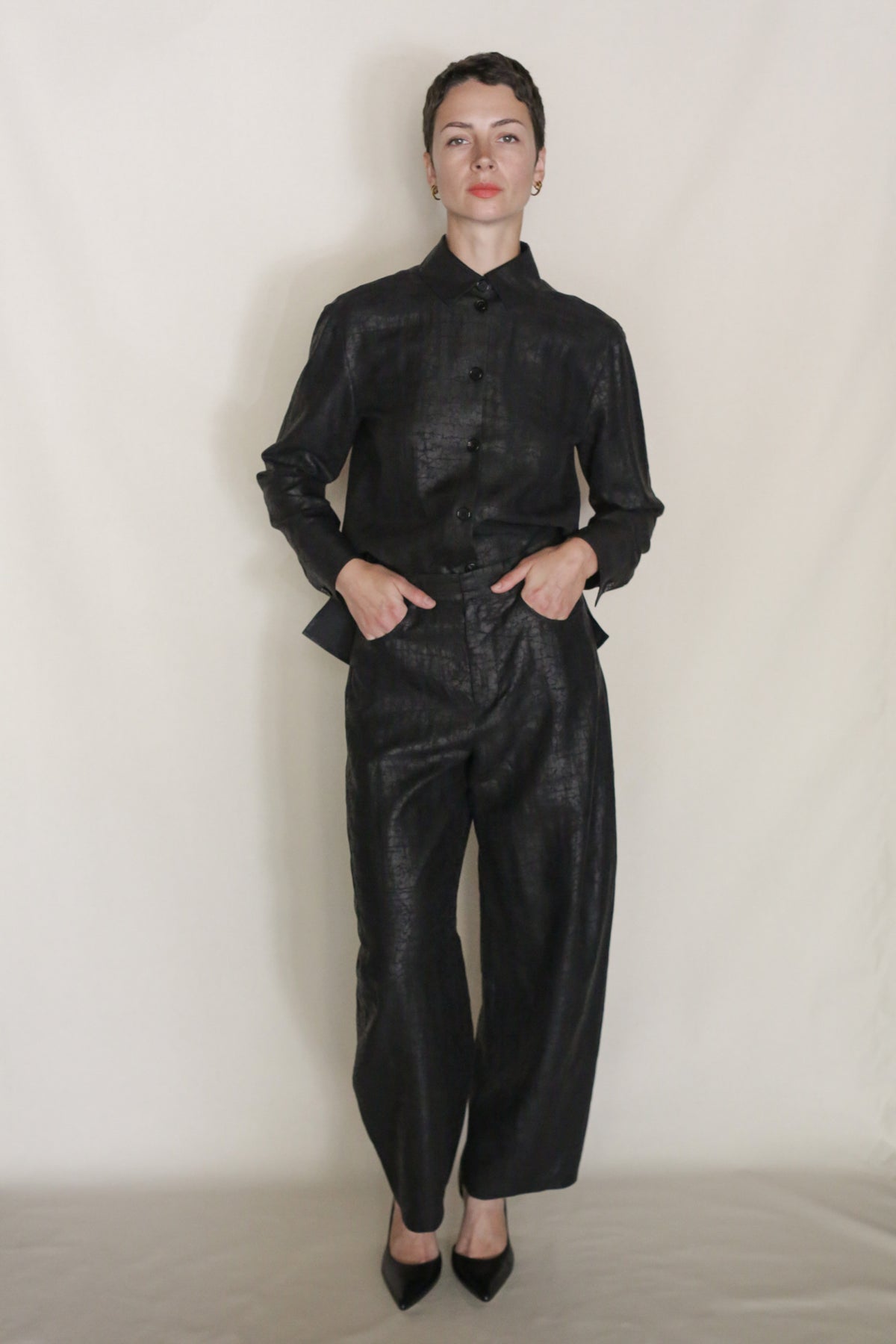 A person with short hair stands against a plain light background, wearing black textured button-up shirt and Earth Mark Barrel Trousers for a modern silhouette. Their hands are in their pockets as they look at the camera.