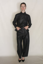Load image into Gallery viewer, A person with short dark hair and a neutral expression stands against a plain light background, wearing shiny black Barrel Leg Trousers | Xiang Yun Silk Earth Mark, a matching button-up shirt, and black pointed-toe heels, hands in pockets.