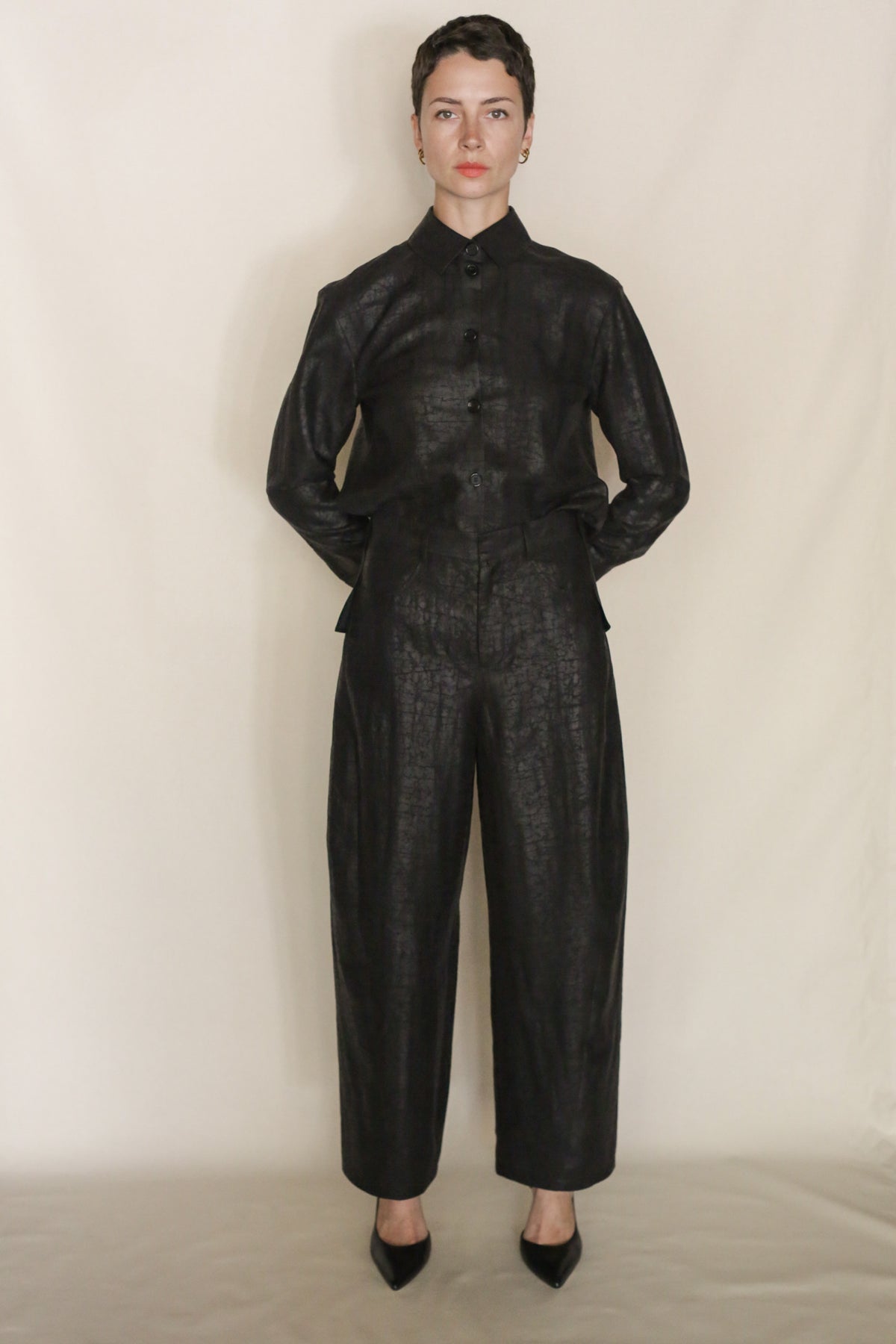 A person with short dark hair stands against a plain light background, wearing a black textured Xiang Yun Silk button-up long-sleeve top and matching Earth Mark Barrel Trousers with black pointed shoes, looking directly at the camera.