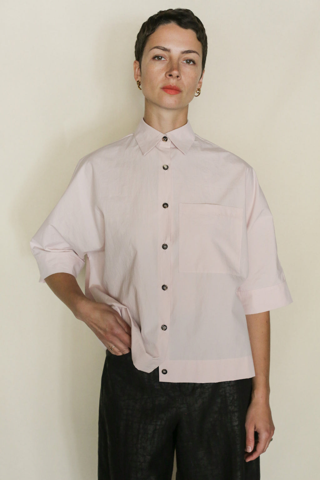Person with short dark hair wears the Work Shirt Crop | Blush—an organic typewriter cotton loose light pink cropped shirt with a large front pocket—paired with dark textured pants. Handmade in San Francisco, shown against a plain light background.