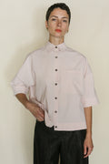 Person with short dark hair wears the Work Shirt Crop | Blush—an organic typewriter cotton loose light pink cropped shirt with a large front pocket—paired with dark textured pants. Handmade in San Francisco, shown against a plain light background.