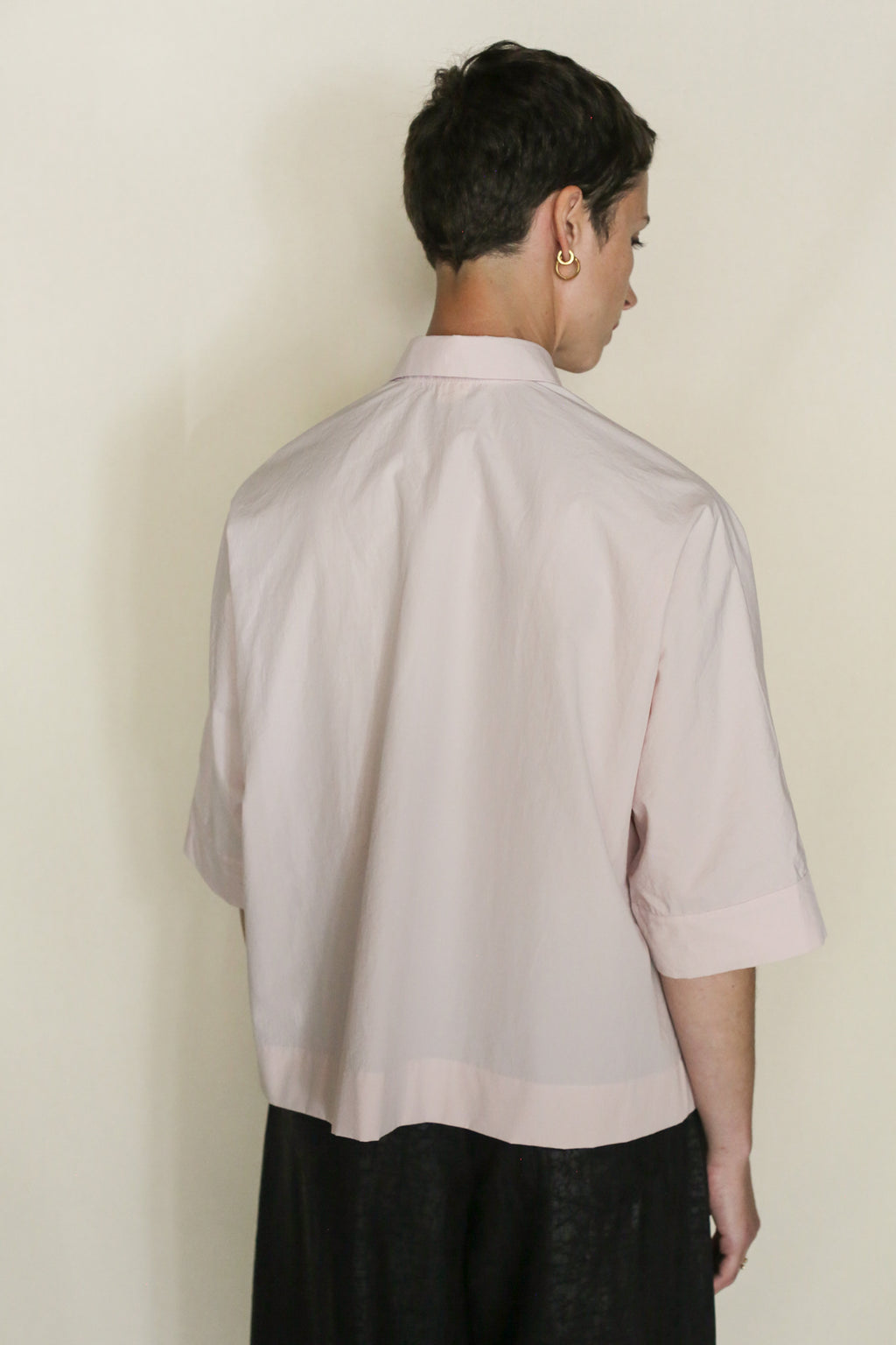 A person with short brown hair, wearing the Work Shirt Crop | Blush and black organic typewriter cotton pants, stands facing away from the camera against a plain light background.