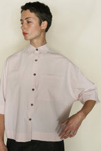 Load image into Gallery viewer, A person with short dark hair wears the Work Shirt Crop in Organic Typewriter Cotton Blush, featuring a cropped fit, front pocket, and dark buttons. They look to the side with one arm bent.