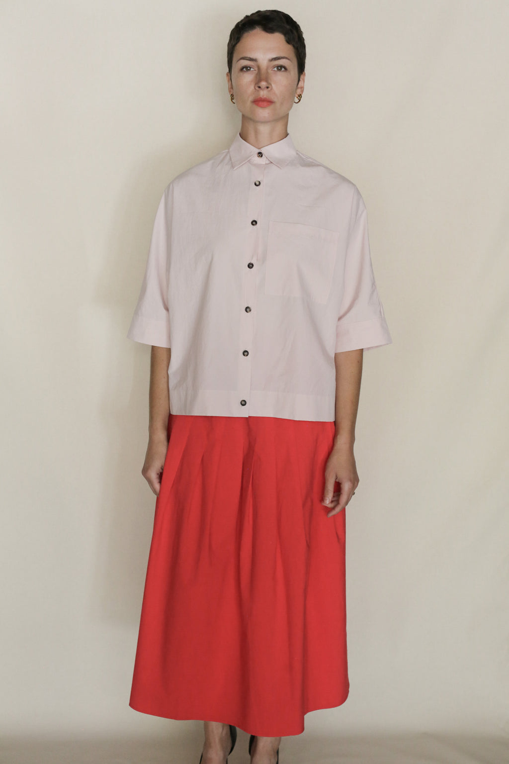 Person wearing the Work Shirt Crop | Blush, made from organic typewriter cotton, paired with a high-waisted bright red skirt—handmade in San Francisco—stands against a plain beige background.