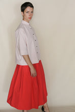 Load image into Gallery viewer, A person with short dark hair wears the Work Shirt Crop in Organic Typewriter Cotton Blush, paired with a bright red full midi skirt, standing in profile against a plain beige background.