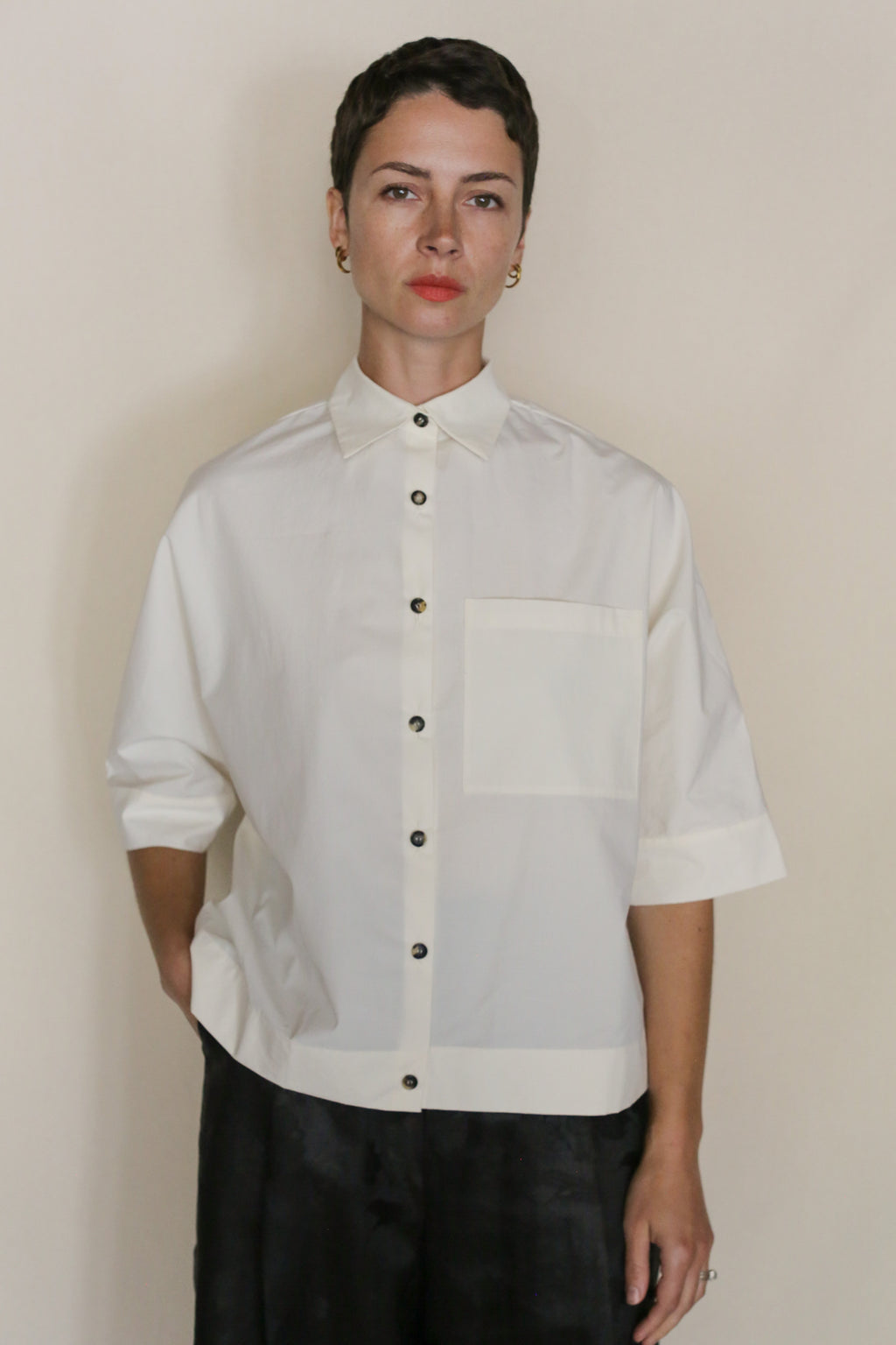A person with short dark hair wears the Work Shirt Crop | Undyed, a handmade cropped shirt crafted from organic typewriter cotton with short sleeves and a large chest pocket, standing against a neutral background.